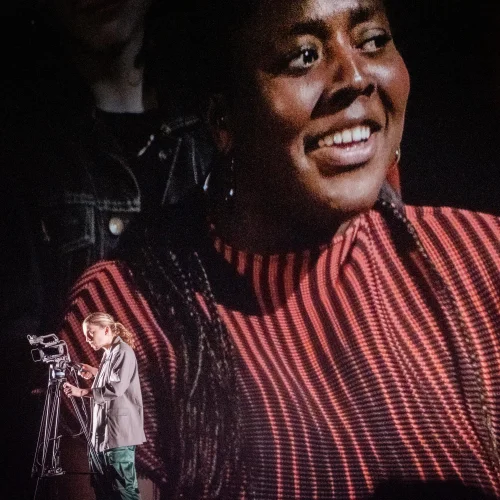 A woman with a camera is in the corner of the image whilst a large projection of another woman is behind her.