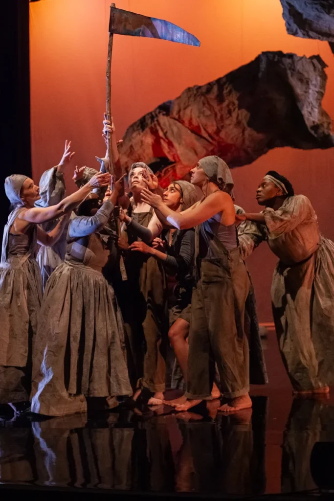Group of witches in flowing grey dresses surrounding a penant flag on a stave